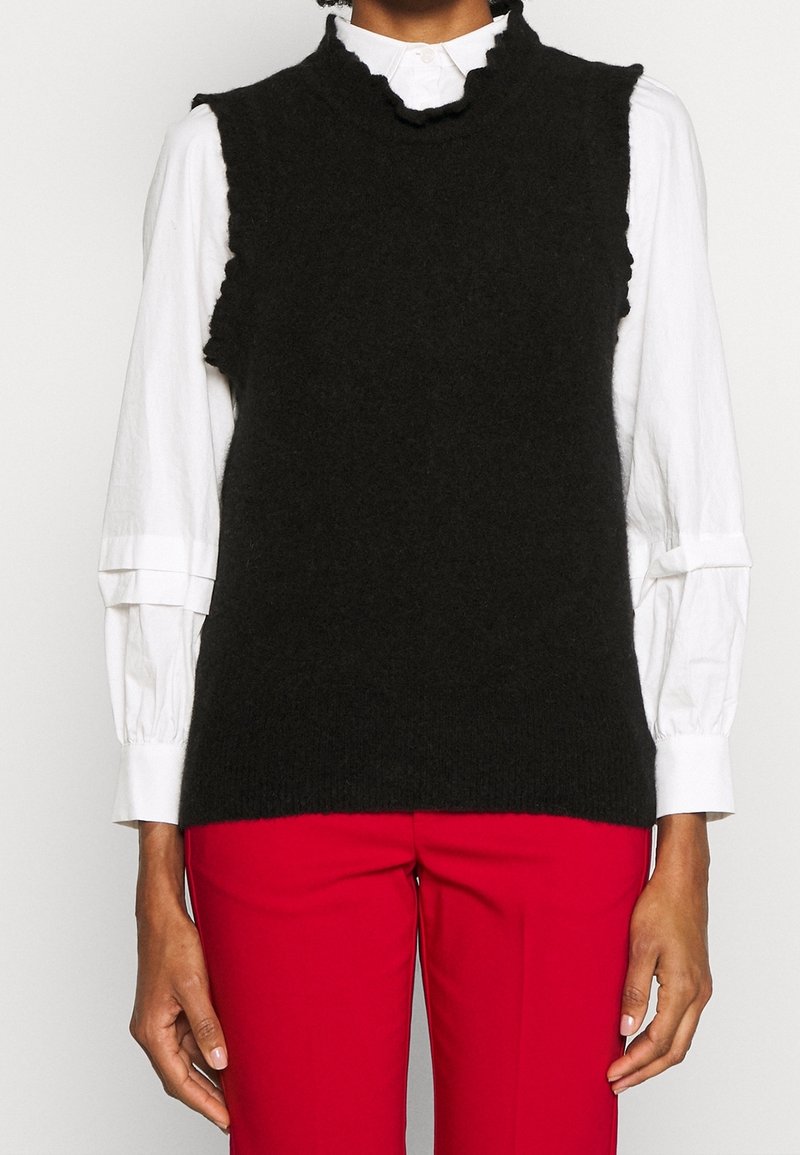 Personne portant un gilet en maille noir sans manches sur une chemise blanche à manches longues avec des poignets volantés et un pantalon rouge.