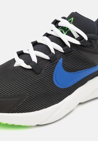 Zwarte atletische schoen met getextureerde bovenkant, blauwe Nike swoosh, witte veters en groene accenten op de zool. Lichtgewicht ontwerp voor optimale prestaties.