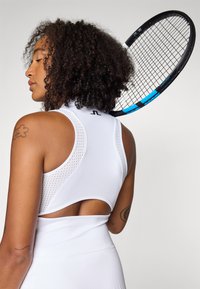 Vit sport-bh med mesh-design, öppen rygg och logodetalj. En tenniracket vilar på axeln i bakgrunden.