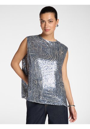 Frau trägt ein ärmelloses silber-blaues Paillettenoberteil mit geometrischen Mustern, dunkle Hosen, silberne Ohrringe und ein silbernes Armband.