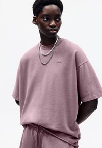 Losse roze katoenen t-shirt met een geribbelde halslijn en een klein logo, gecombineerd met een zilveren en parelachtige gelaagd ketting.
