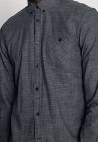 Grijze button-up shirt van textiele stof, met een gestructureerde kraag, zes zwarte knopen en één borstzak.