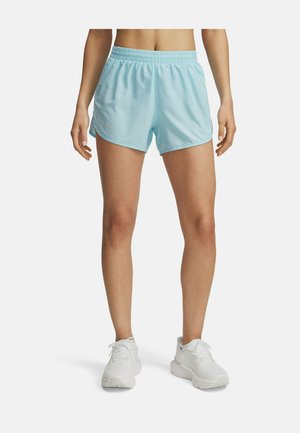 Kvinna som bär ljusblå träningsshorts och vita löparskor, står med benen något isär mot en vit bakgrund.
