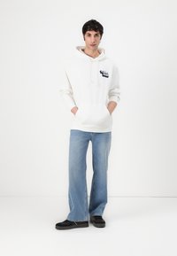 Hoodie blanche avec une poche avant et un logo, associé à un jean évasé bleu clair et des baskets noires. Tissu lisse et coupe décontractée.