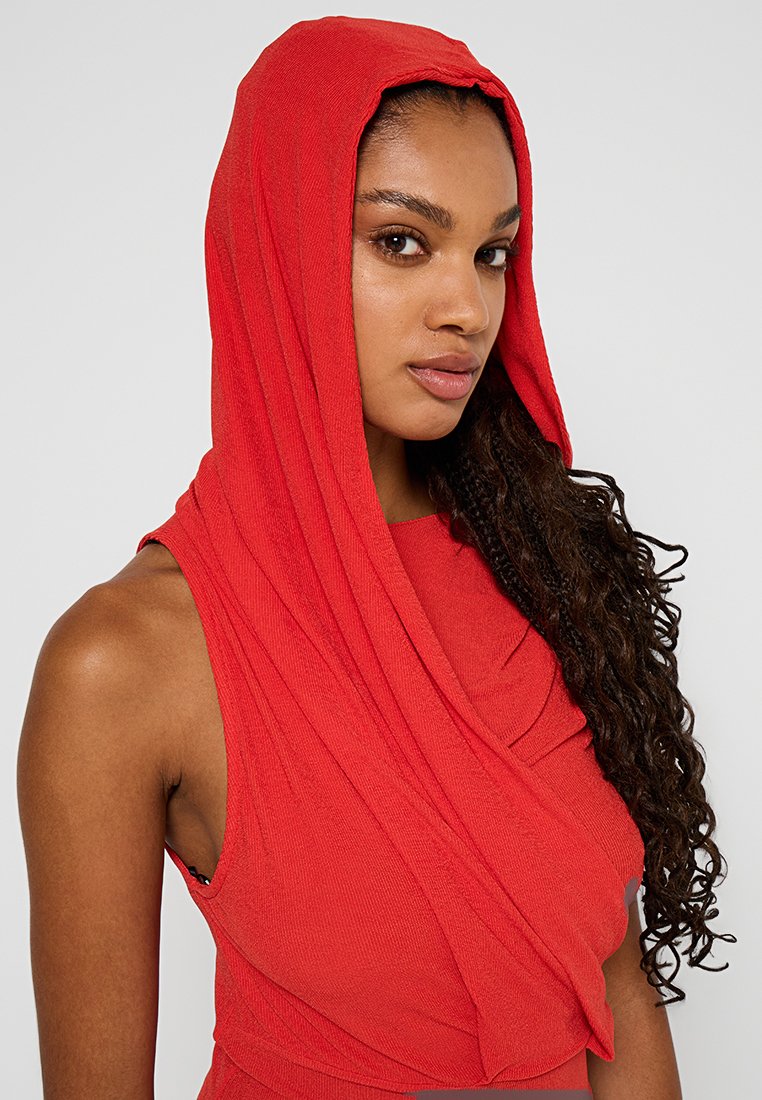 Czerwona bezrękawnik z drapowanym kapturem, z prążkowaną teksturą i miękkim materiałem. Zbliżenie na twarz modelki i ramię.