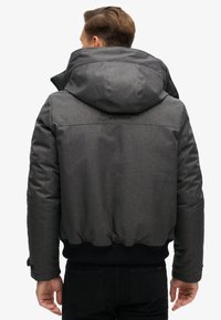 Grijze pufferjack met een capuchon, voorzien van een gestructureerde stof, ribgebreide manchetten en een elastische zoom. Logo op de rug nabij de halslijn.