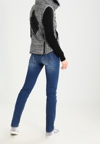 Grå og sort mønstret ærmeløs vest med lynlås og faux fur krave, lagdelt over en sort striktrøje, parret med blå jeans og sølvfarvede sneakers.