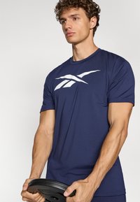 Námornícky modré atletické tričko s bielym logom na hrudi, krátke rukávy a priliehavý strih, držené s činilom v rukách.