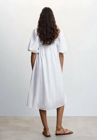 Weißes Baumwollkleid mit Puffärmeln, gerafftem Taillenbereich und einer Länge bis zur Mitte der Wade. Es hat eine glatte Textur und ein einfaches, elegantes Design. Braune Sandalen.