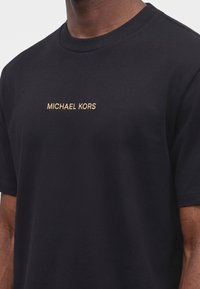 Man wearing black crew neck t-shirt with "MICHAEL KORS" embroidered in gold thread across the chest.