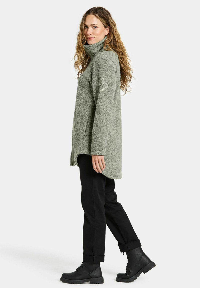 Grüner Fleece-Pullover mit hohem Kragen und strukturierter Oberfläche. Kombiniert mit schwarzen Hosen und robusten schwarzen Stiefeln.