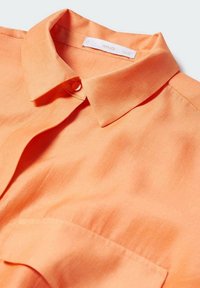 Camicia a maniche corte in tessuto arancione brillante con una texture liscia, colletto classico e una tasca sul petto. Presenta una chiusura con bottoni.