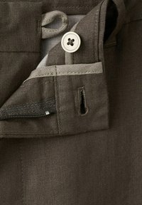 Prim-plan al taliei pantalonilor maro din in, arătând nasturele alb, găurelele pentru nasture, fermoarul și detaliul buclei din materialul interior.