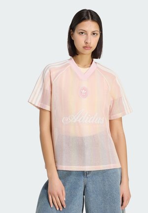 Młoda kobieta o prostych ciemnych włosach, ubrana w różową siateczkową koszulkę Adidas z białymi paskami oraz niebieskie dżinsy, stojąca na tle jednolitej ściany.