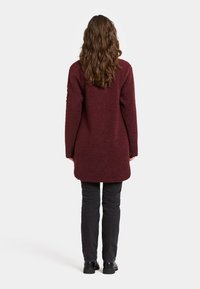 Bordeaux pitkähihaisessa takissa on tekstuuriltaan fleece-tyyppinen kangas; polven pituinen ja pyöristetty helma; yhdistetty mustiin housuihin ja mustiin jalkineisiin.
