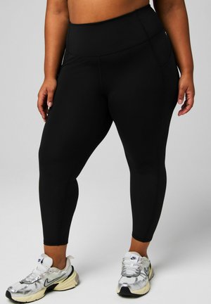 High-waisted black leggings made of stretchy fabric, featuring a smooth texture and side pocket, paired with silver and white athletic shoes.