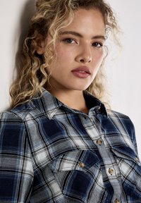 Niebieska i biała koszula w kratę z zapięciem na guziki, posiadająca dwie przednie kieszenie i kołnierzyk. Wykonana z tkaniny o miękkiej teksturze.