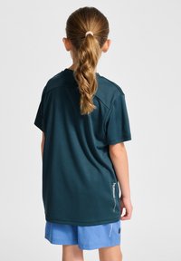 Mörk teal, kortärmad tröja i jersey med en jämn textur. Har en rundad halsringning och en vit logodetalj på sidan, tillsammans med ljusblå shorts.