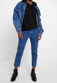 Chaqueta de mezclilla con lavado azul claro, sudadera con capucha negra, jeans azul ajustados con raya lateral y botas negras hasta el tobillo.