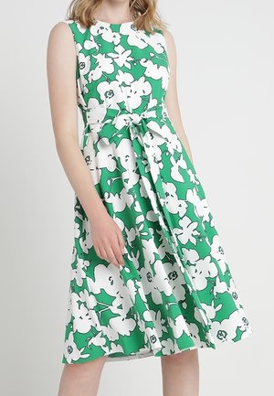 Ærmeløs grøn kjole med stort hvidt blomstermønster og et bælte bundet om taljen, båret af en stående person mod en ensfarvet baggrund.