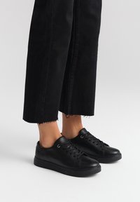 Sneakers neri a basso profilo realizzati in pelle liscia, con punta rotonda, lacci frontali e suola piatta in gomma. Abbinati a pantaloni neri a 3/4.
