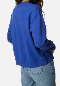 Mujer vestida con un sudadera azul lisa y jeans azul claros, mostrada de espaldas contra un fondo blanco.