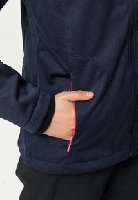 Chaqueta softshell en color navy con textura suave, que presenta un detalle en forma de cremallera rosa en el bolsillo y un diseño entallado para un ajuste perfecto.