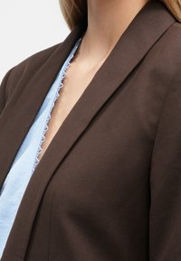 Brązowa marynarka z wyciętym kołnierzem i czystymi liniami, zestawiona z jasnoniebieską bluzką z detalem w postaci karbowanego dekoltu. Miękka, teksturowana tkanina.