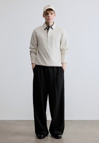 Beige sweatshirt med en struktureret krave, sorte bukser og en lysebrun kasket med tekst. Glatte teksturer og afslappet pasform.