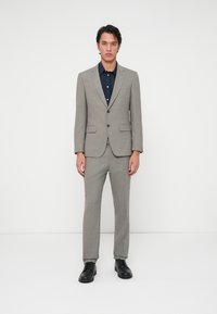 Ανοιχτό γκρι κοστούμι με λεπτή υφή, κοντή εφαρμογή, σακάκι με δύο κουμπιά, ταιριαστά παντελόνια και σκούρο μπλε πουκάμισο. Μαύρες δερμάτινες παπούτσια με κορδόνια.