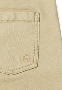 Tejido de mezclilla beige con un bolsillo trasero cosido y un pequeño logotipo bordado cerca de la esquina inferior derecha del bolsillo.