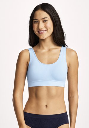 Junge Frau mit langen dunklen Haaren lächelt, trägt einen hellblauen Sport-BH und dunkle gemusterte Unterwäsche vor einem schlichten hellen Hintergrund.