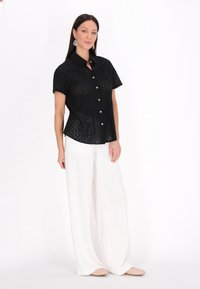 Camicia nera a maniche corte con bottoni e motivo a occhiello, abbinata a pantaloni bianchi larghi. Leggera e dal taglio rilassato.