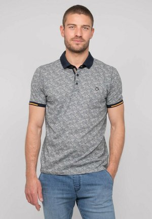 Homme aux cheveux courts et barbe portant un polo gris à motifs avec un col noir et un jean bleu, debout devant un fond neutre.