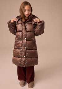 Manteau matelassé marron avec capuche, orné de boutons dorés et de grandes poches. Porté avec un pantalon large bordeaux foncé et des bottes beiges.
