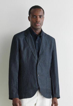 Mann trägt einen dunklen Nadelstreifen-Blazer über einem schwarzen Hemd und weißen Hosen, steht an einer schlichten hellgrauen Wand.