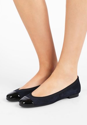 Pieds portant des ballerines noires avec des bouts brillants et un corps en daim, présentés sur fond blanc.