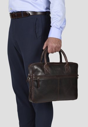 Brown leather briefcase with double handles, front zipper pocket, and textured surface. Worn in hand next to navy trousers.
