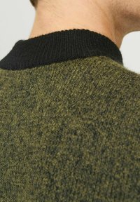 Knitted sweater with a textured olive green body and a thick black ribbed collar, focusing on the material's warmth and tactile quality.