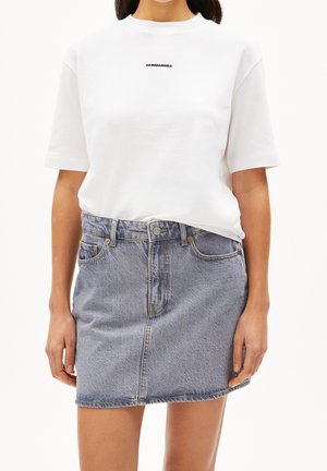 Mujer que lleva una camiseta blanca de manga corta con texto pequeño en negro y una falda vaquera azul claro con bolsillos delanteros y cierre de botones.
