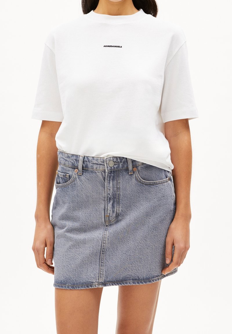 Femme portant un T-shirt blanc à manches courtes avec un petit texte noir et une jupe en jean bleu clair avec poches avant et fermeture par boutons.