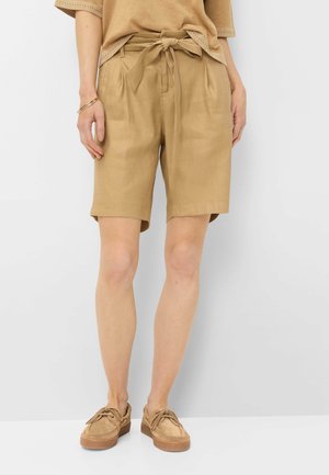 Person iført beige knælange shorts med et bundet bælte i taljen, kombineret med matchende beige sko og en lysebrun T-shirt med korte ærmer.