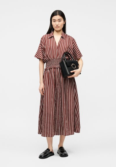 Γυναίκα με μίντι φόρεμα με κάθετες καφέ και λευκές ρίγες και με ελαστική μέση, κρατάει μαύρη τσάντα και φοράει μαύρα παπούτσια τύπου slip-on.