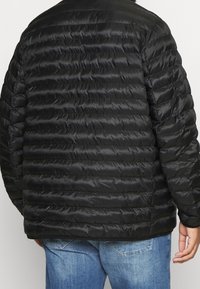 Schwarze Pufferjacke mit einem gesteppten Design. Verfügt über eine weiche, glänzende Oberfläche und eine lockere Passform, kombiniert mit Jeans.