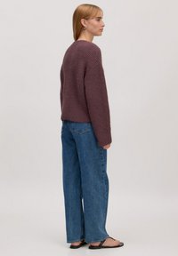 Trui in donker mauve breiwerk met een cropped, boxy fit. Gecombineerd met brede blauwe spijkerbroek. Model staat met haar rug naar de camera.