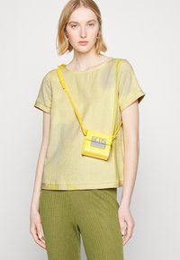 Top amarelo com padrão de manga curta e decote redondo, combinado com calças texturizadas verdes com costelas e uma pequena mala tiracolo amarela com um logotipo.