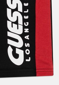 Čierna tkanina s červeným bočným panelom. Má tučný biely nápis "GUESS LOS ANGELES" na čiernej. Mäkká bavlnená textúra; neformálny dizajn.