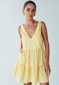 Geltona medvilninė suknelė su gilia V formos iškirpte ir sluoksniuotu raukinio dizainu. Turi platų petnešėlių ir kišenes abiejose pusėse. Lygi tekstūra.