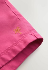 Tissu rose vif à texture lisse, présentant un petit palmier brodé en or sur l'ourlet, mettant en valeur des éléments de design simples.