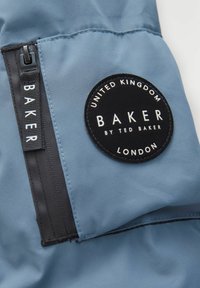 Mėlynas audinys su lygiu paviršiumi, turintis juodą apvalų etiketę su užrašu "BAKER by Ted Baker" ir juodą užtrauktuką.